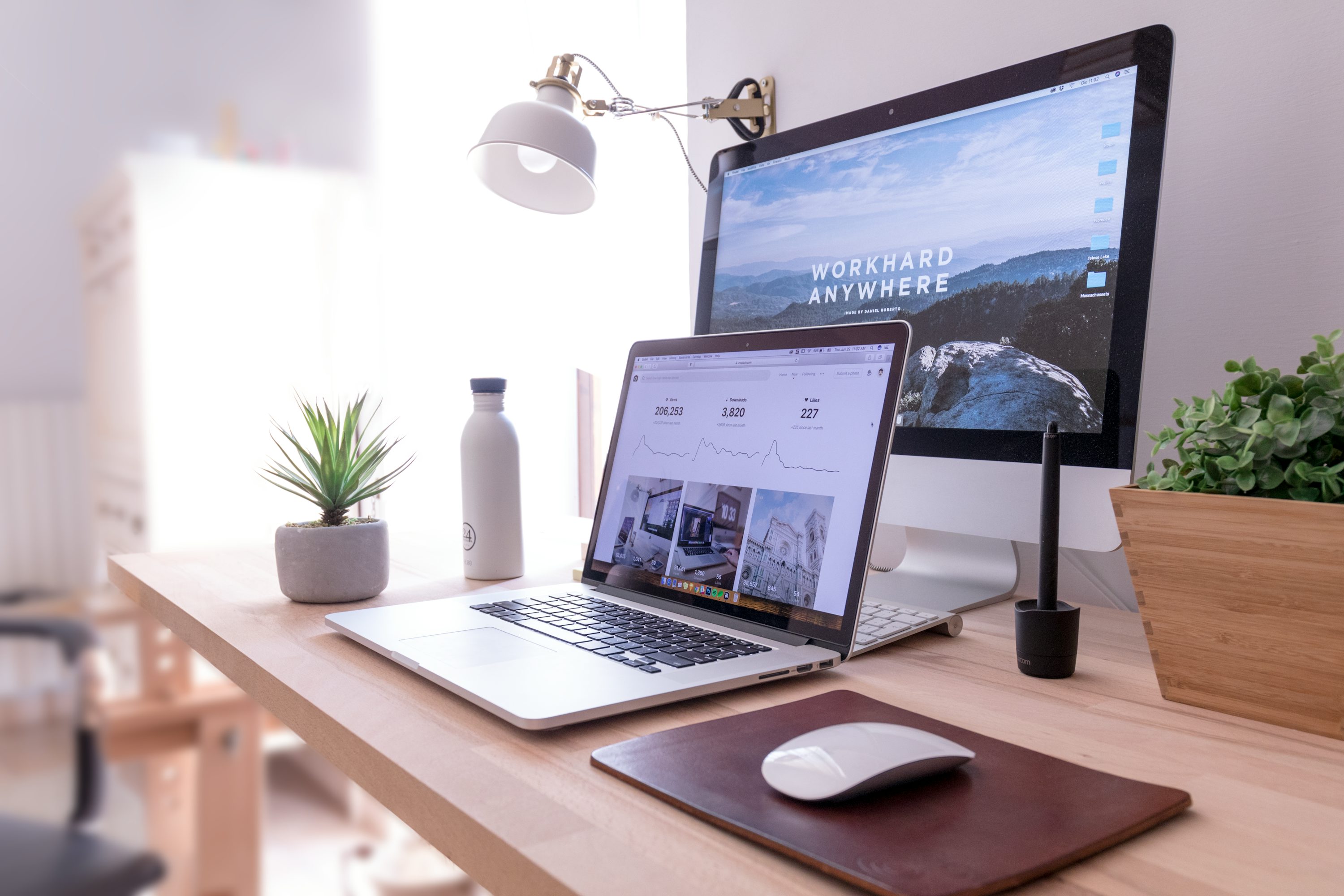 Poste de travail moderne avec ordinateur portable et écran desktop affichant un site web, lampe, plante et souris sur une table en bois.