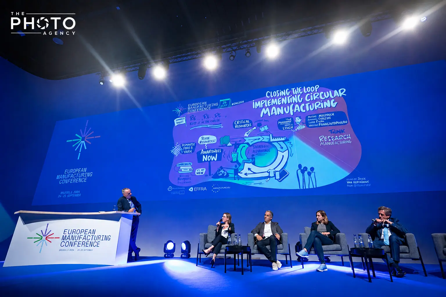 Conférence européenne sur la fabrication, avec un podium et panneaux bleus, orateurs assis sur scène et une grande présentation graphique illuminée. Conférence européenne sur la fabrication, avec un podium et panneaux bleus, orateurs assis sur scène et une grande présentation graphique illuminée.