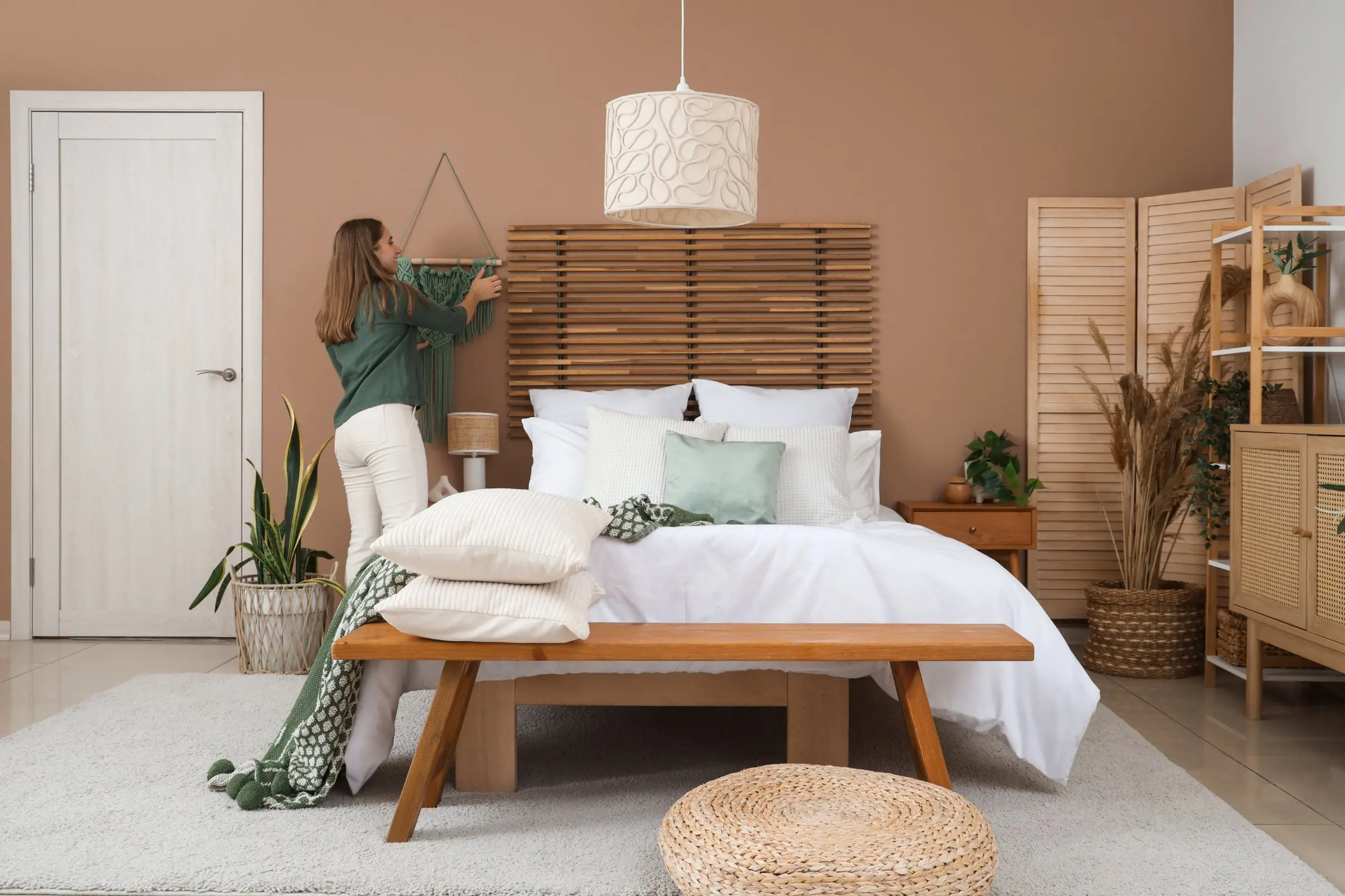 Salle de lit professionnelle avec lit confortable, tête de lit en bois, linge blanc et coussins pastel; vendeuse ajustant une housse sur le mur. Salle de lit professionnelle avec lit confortable, tête de lit en bois, linge blanc et coussins pastel; vendeuse ajustant une housse sur le mur.