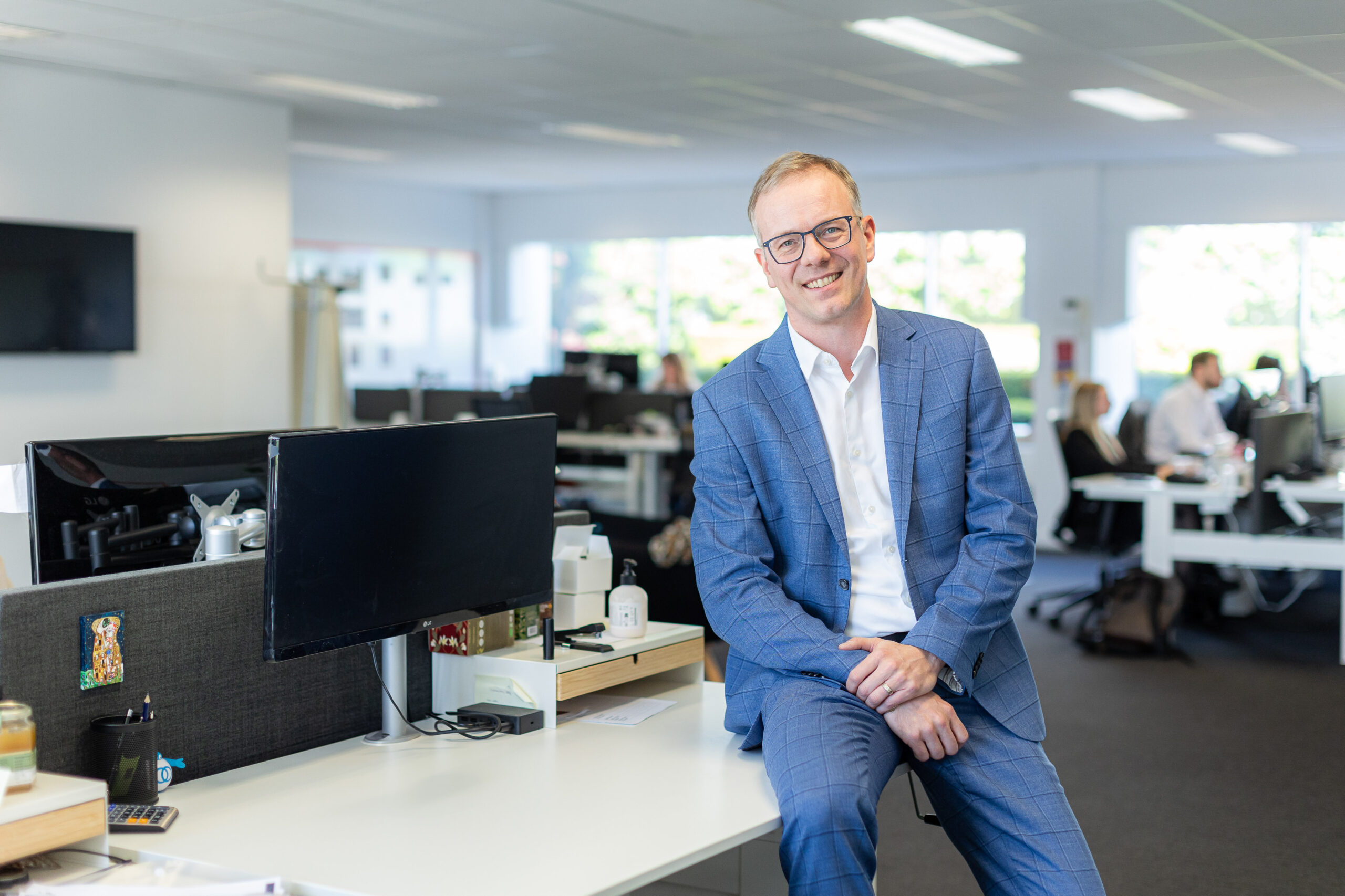 Directeur souriant en bureau ouvert moderne, costume bleu, près d’un poste de travail avec écran et objets de bureau.