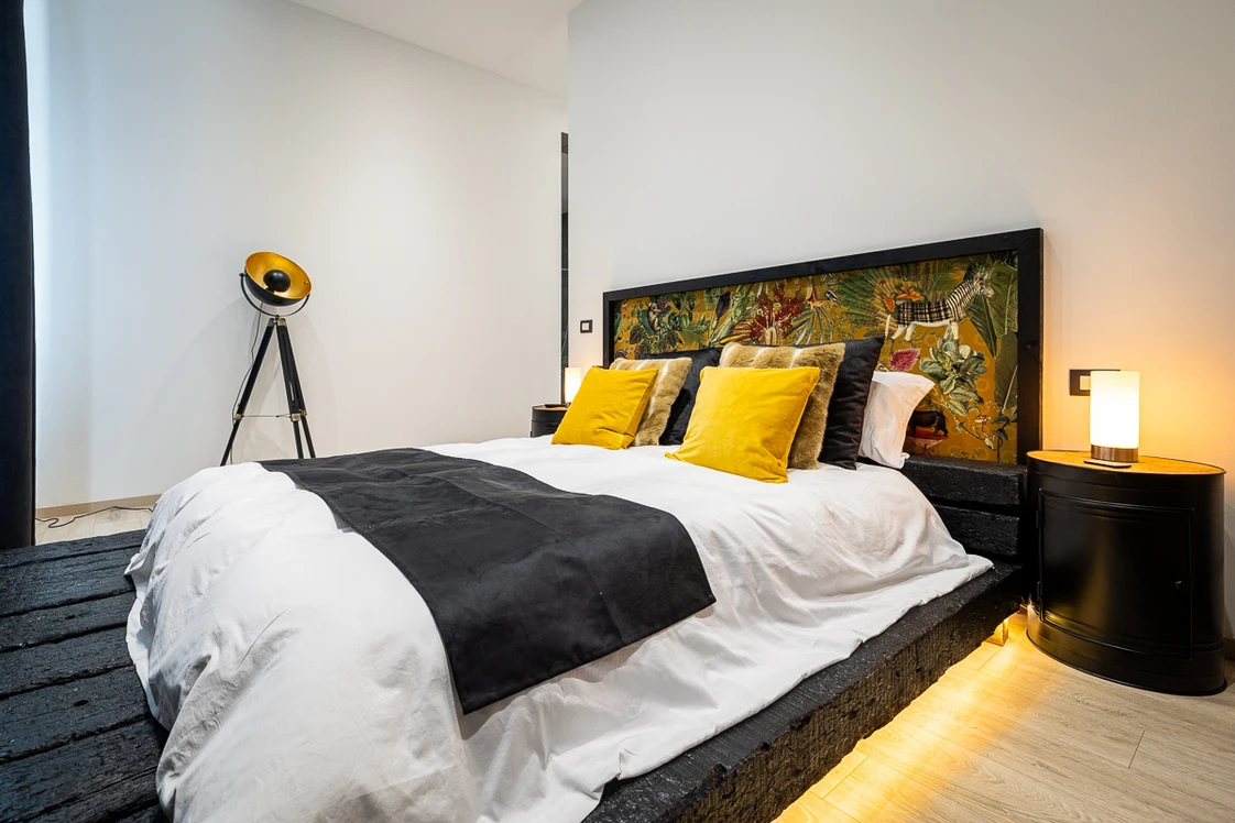 Chambre moderne avec lit king-size, couette blanche et couvre-lit noir, etage surélevé, coussins jaunes et dossier décoratif floral. lampe de chevet et lampadaire industriel dans l’angle.