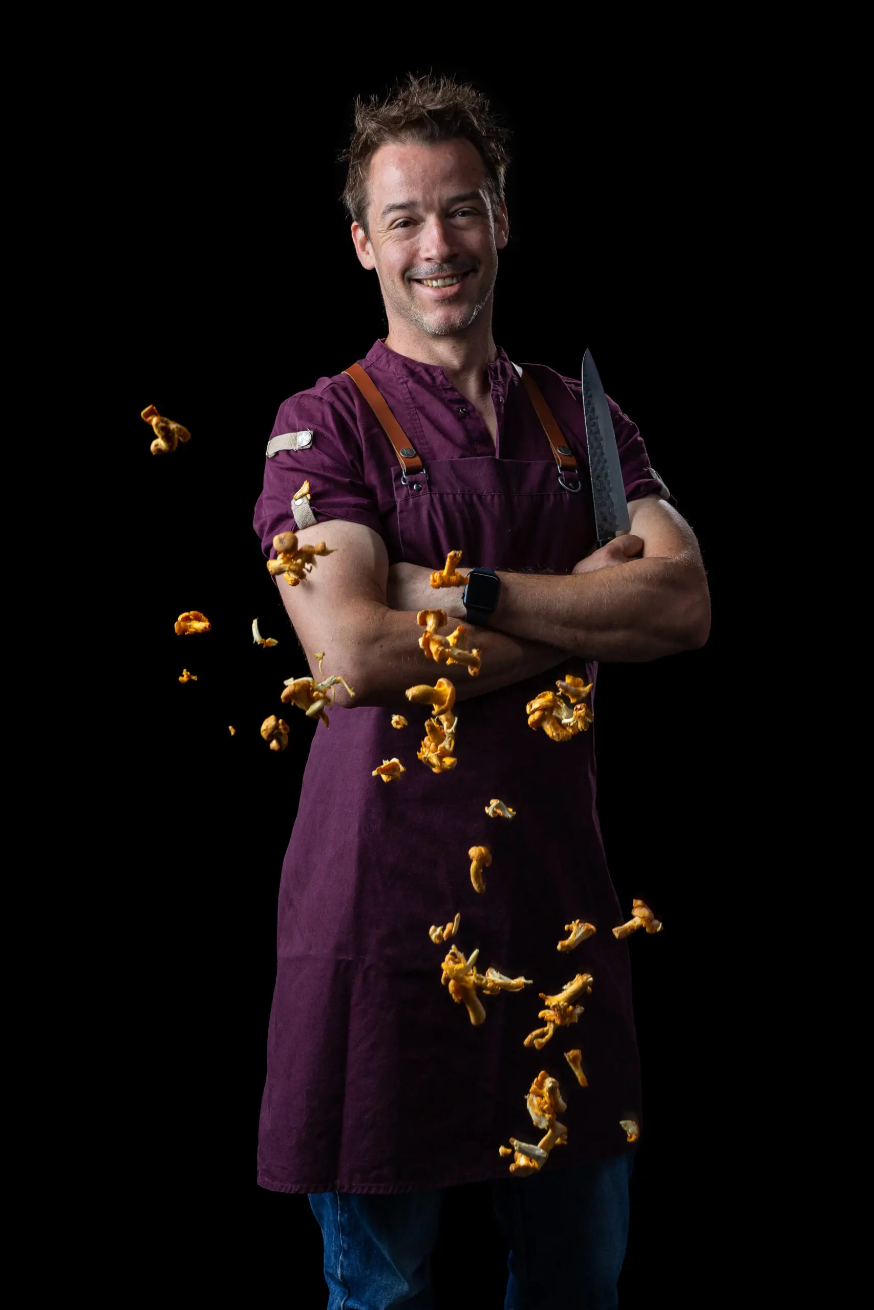 Chef cuisinier en barbeque, portant tablier violet et couteau, avec éclats de popcorn autour sur fond noir. Chef cuisinier en barbeque, portant tablier violet et couteau, avec éclats de popcorn autour sur fond noir.