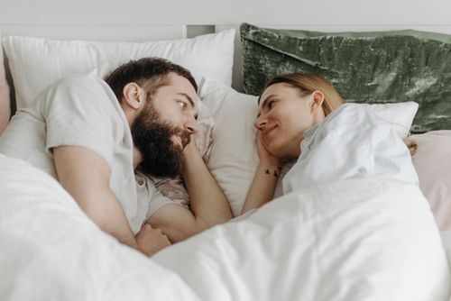 Couple allongé dans un lit, se regardant et souriant sous une grande couette blanche.