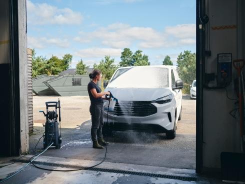 Female operator power-washes a white van at a service bay with hoses and equipment nearby.
