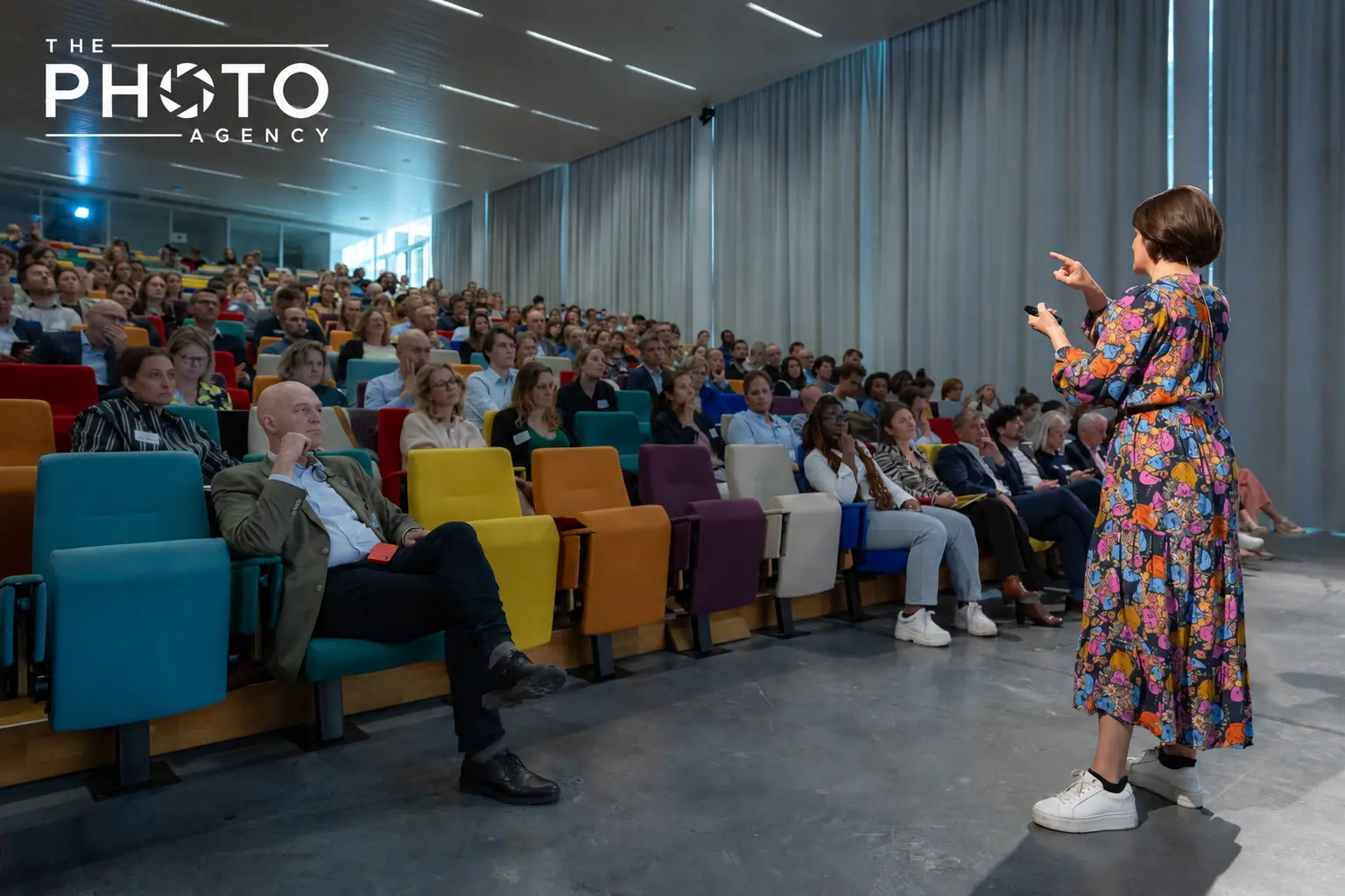 Conférencière en robe à motifs colorés s’adressant à une audience captivée dans une salle de conférence multi-seats colorés, The Photo Agency. Conférencière en robe à motifs colorés s’adressant à une audience captivée dans une salle de conférence multi-seats colorés, The Photo Agency.