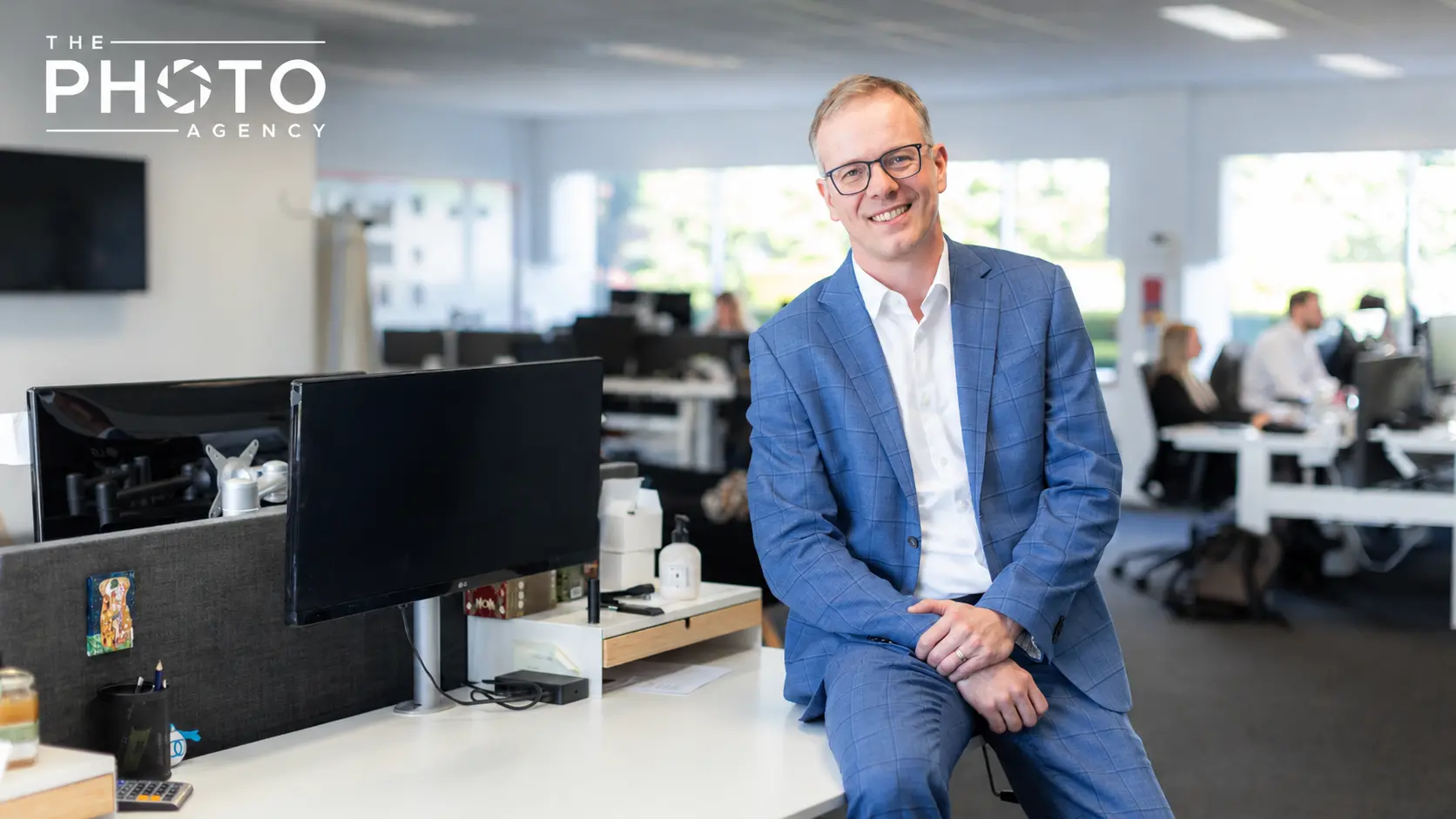 Portrait d’un homme d’affaires en veste bleue dans un open space moderne, assis près d’un bureau avec écran et accessoires. The Photo Agency. Portrait d’un homme d’affaires en veste bleue dans un open space moderne, assis près d’un bureau avec écran et accessoires. The Photo Agency.