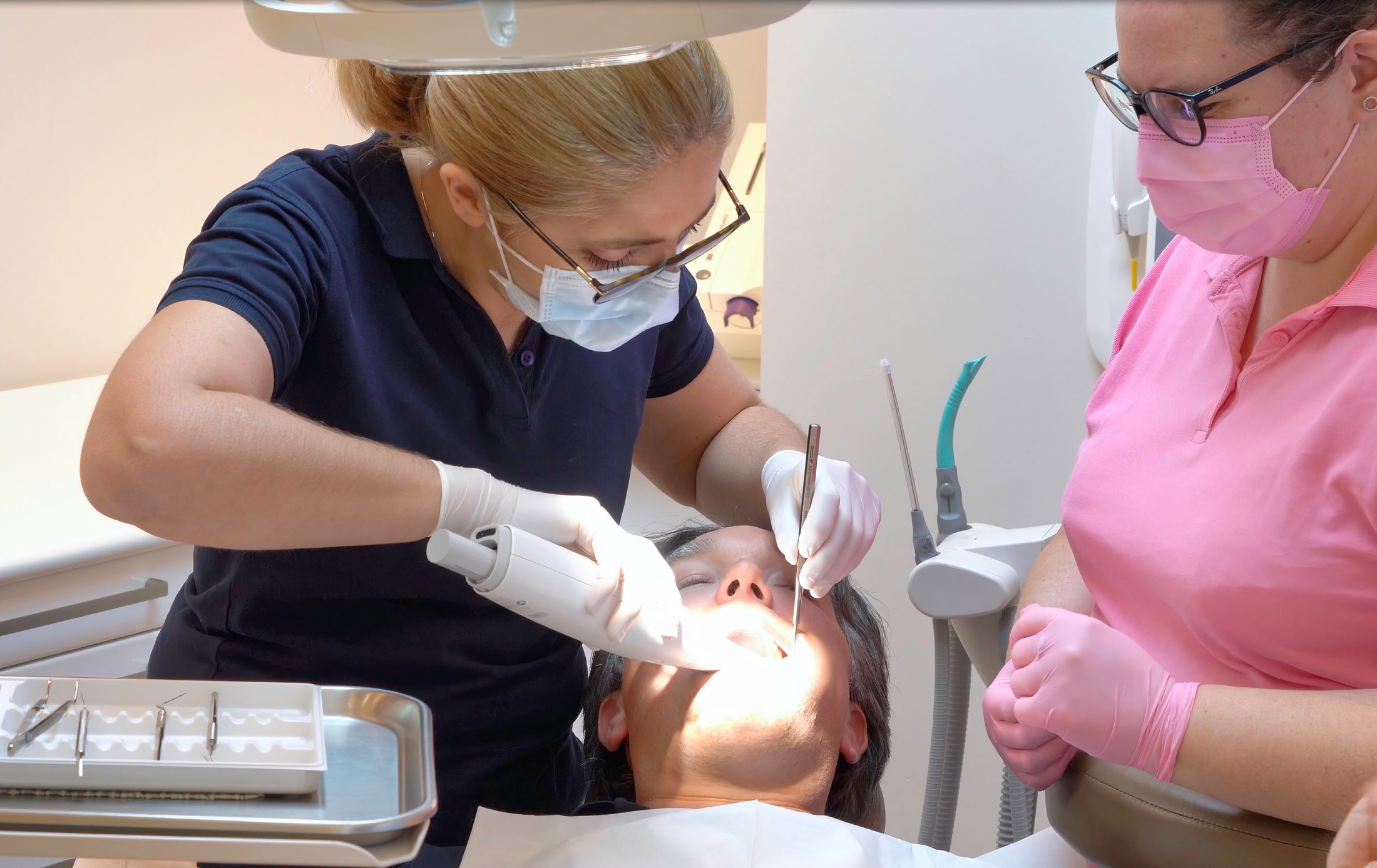 Dentiste en consultation, patient allongé sous lampe opératoire, deux praticiennes portant masque et gants : l’une effectue un soin dentaire avec instrument, l’autre assise à côté prête à intervenir.