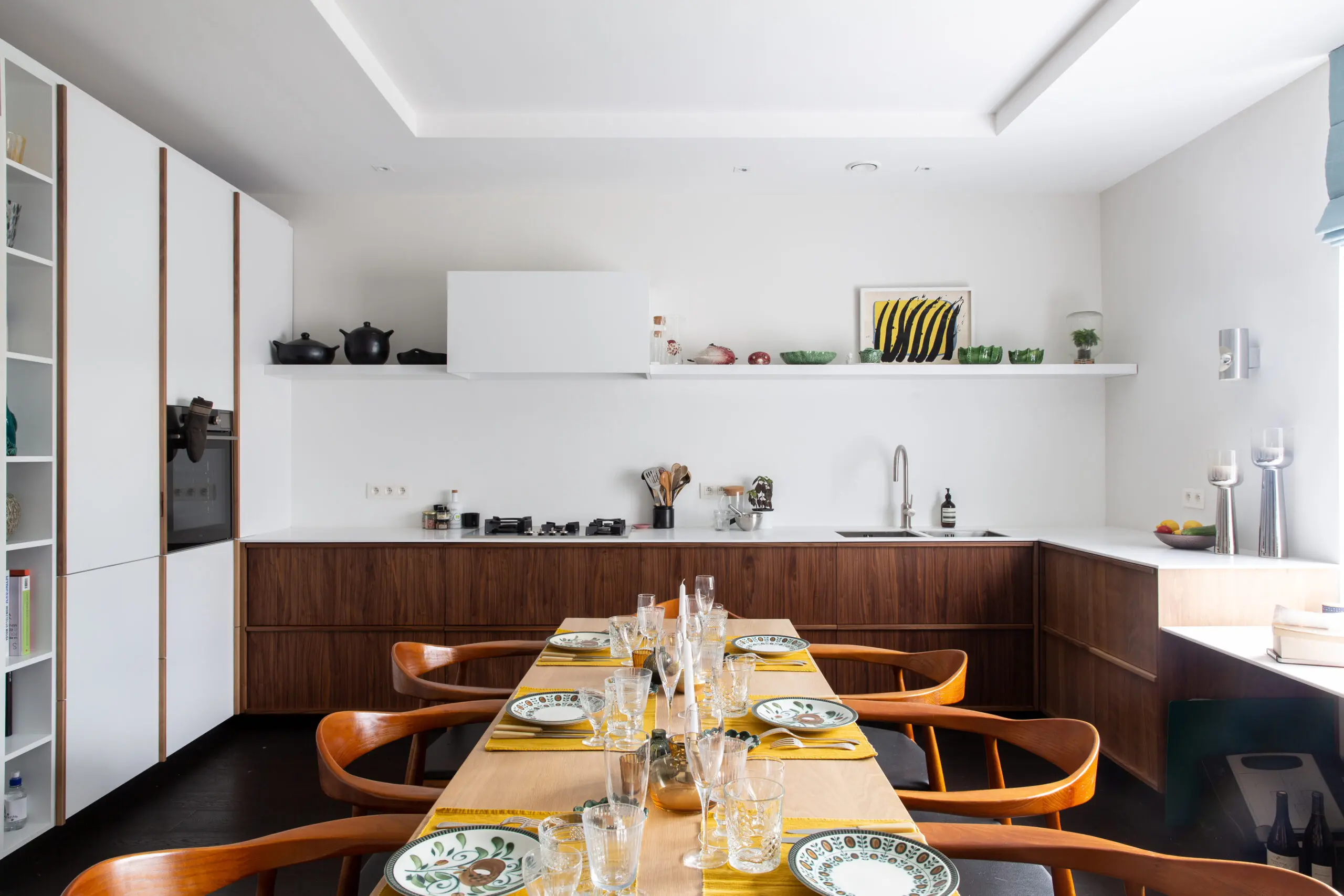 Salle à manger moderne avec long bois et vaisselle colorée, set de table jaune et verres, cuisine ouverte en arrière-plan.