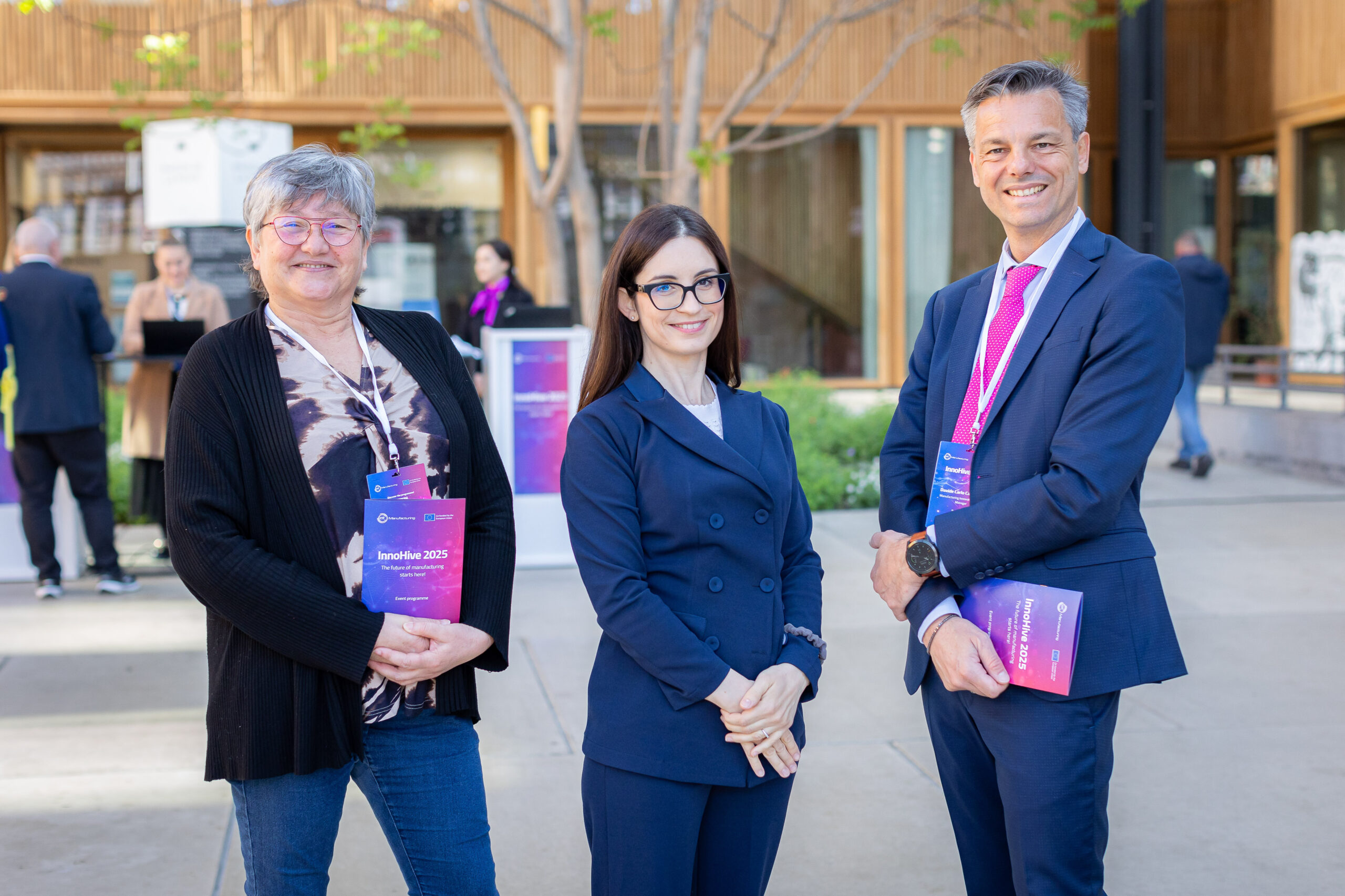 Trois professionnels posent en extérieur lors d’un événement d’entreprise, portant badges et brochures, souriants et élégants.